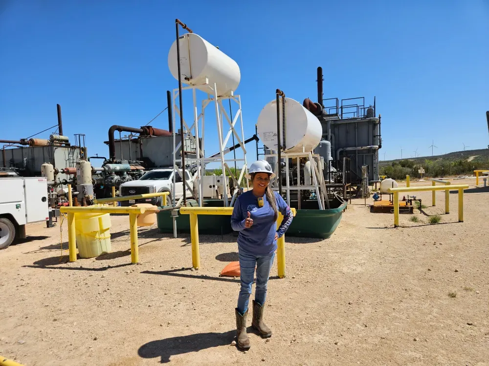 Dorine of Big Fish Rentals and Services, in a hard hat and company shirt, giving a thumbs up on an oil lease site with tank batteries and pump equipment behind her.