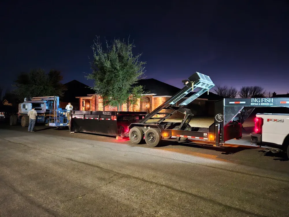 Big Fish Rentals pickup and hook-lift trailer carrying a roll-off container, parked at night in the street outside a lit brick house in Kermit, Texas.
