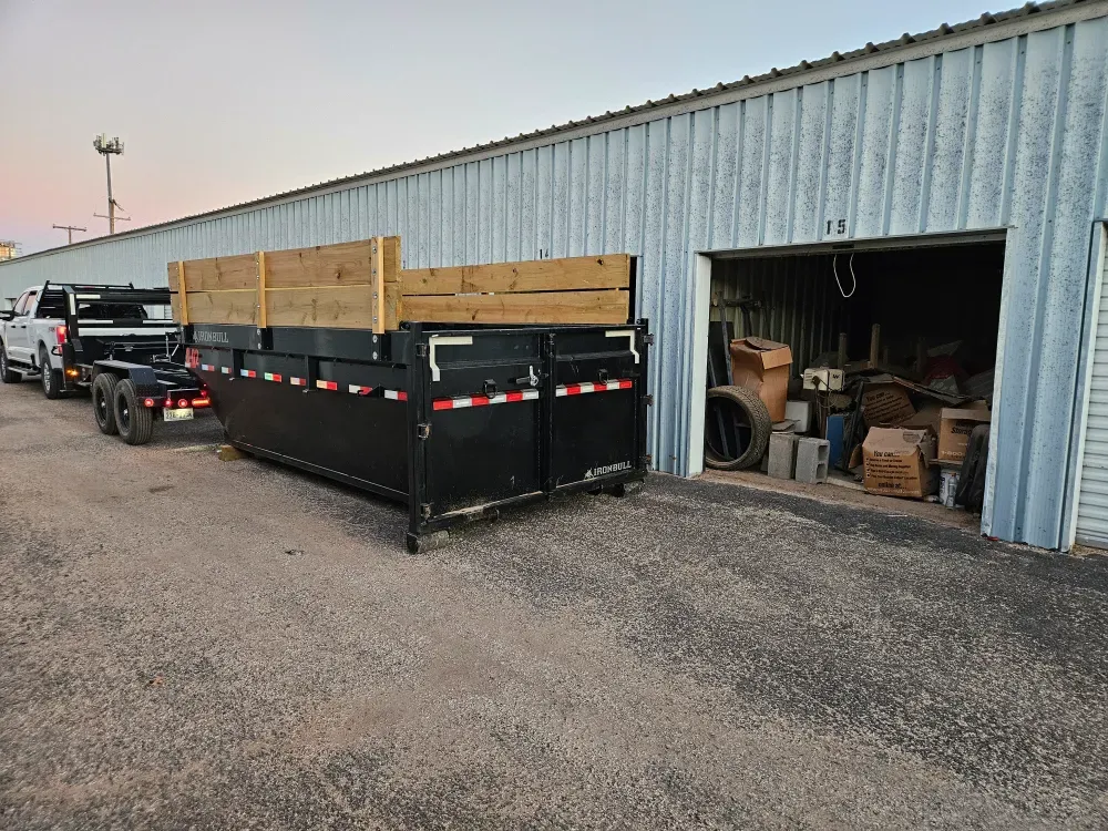 Big Fish roll-off container with wood sideboards parked on gravel beside a row of metal storage units, one unit standing open and packed with boxes and junk, under a pink dusk sky.