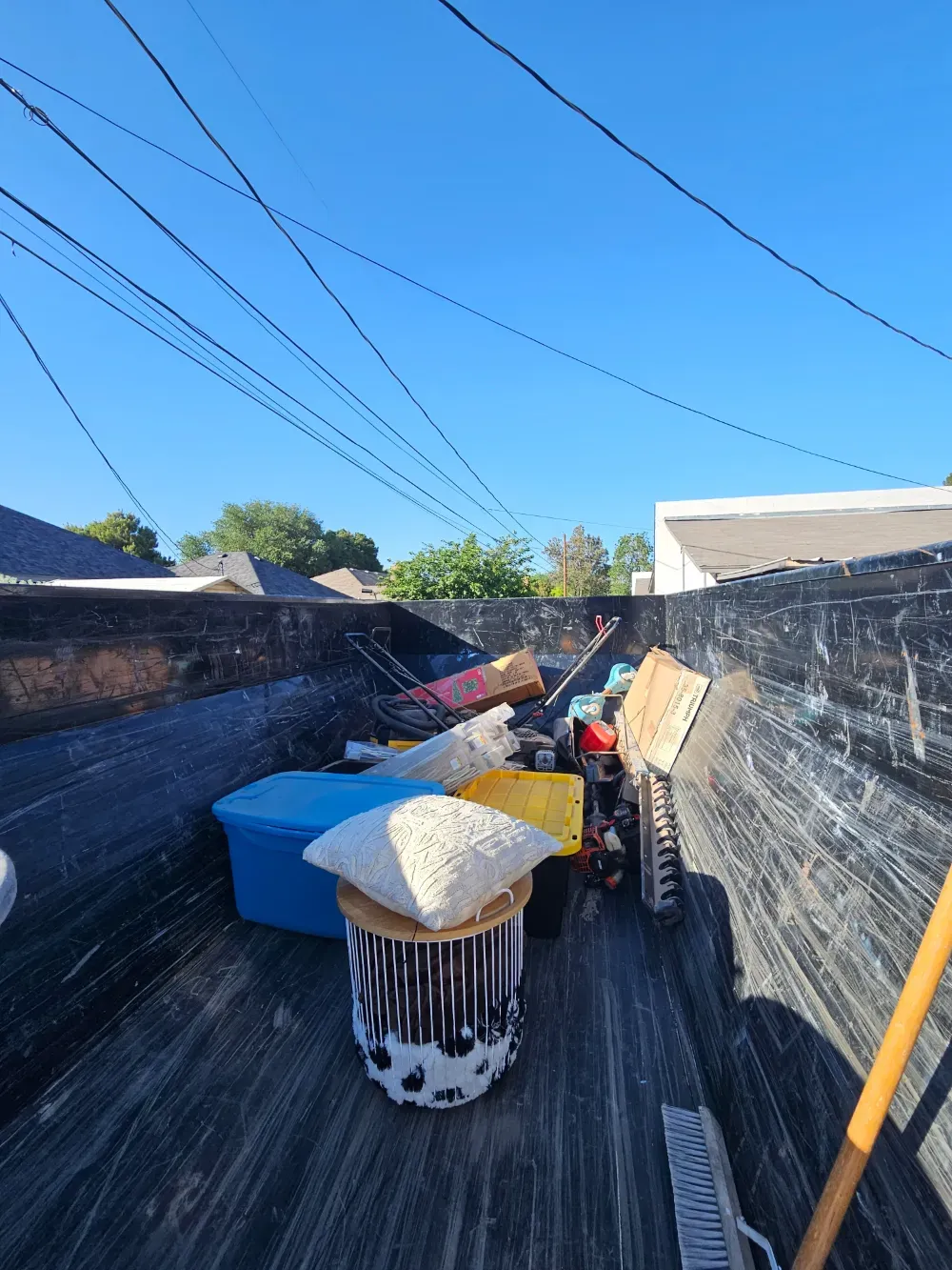 Looking down into a Big Fish Rentals roll-off container from the rim, part-loaded with household items: a blue tote, a laundry hamper, cardboard boxes, a hedge trimmer and a broom.
