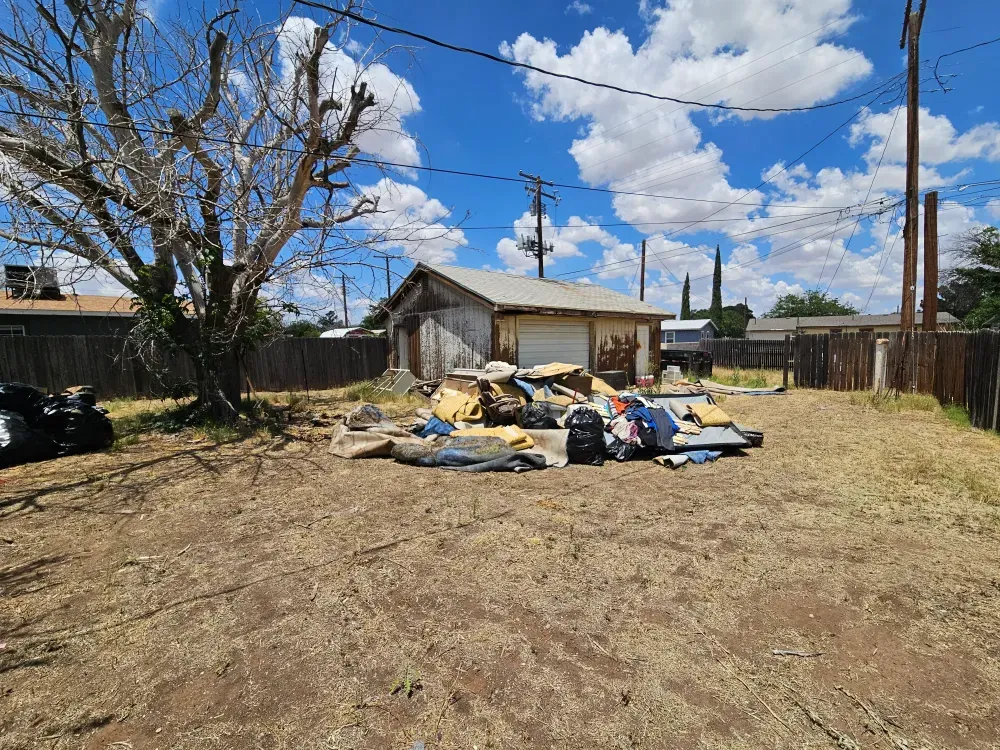 Rolled carpet, cardboard, bagged waste and corrugated panels piled in a back yard beside an old detached garage, the bulky material a city cart will not take.