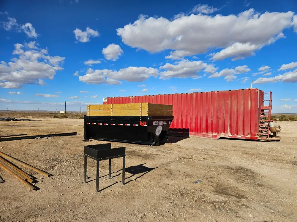 Roll-off container B-02 with timber side extensions set on a caliche lease pad beside a red oilfield tank, flat Permian horizon behind it.