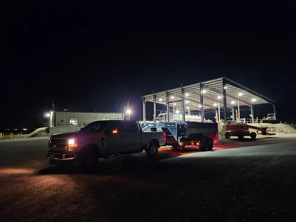 Night on an active build: the Big Fish truck and dump trailer parked on the gravel beside a floodlit steel-frame building under construction.