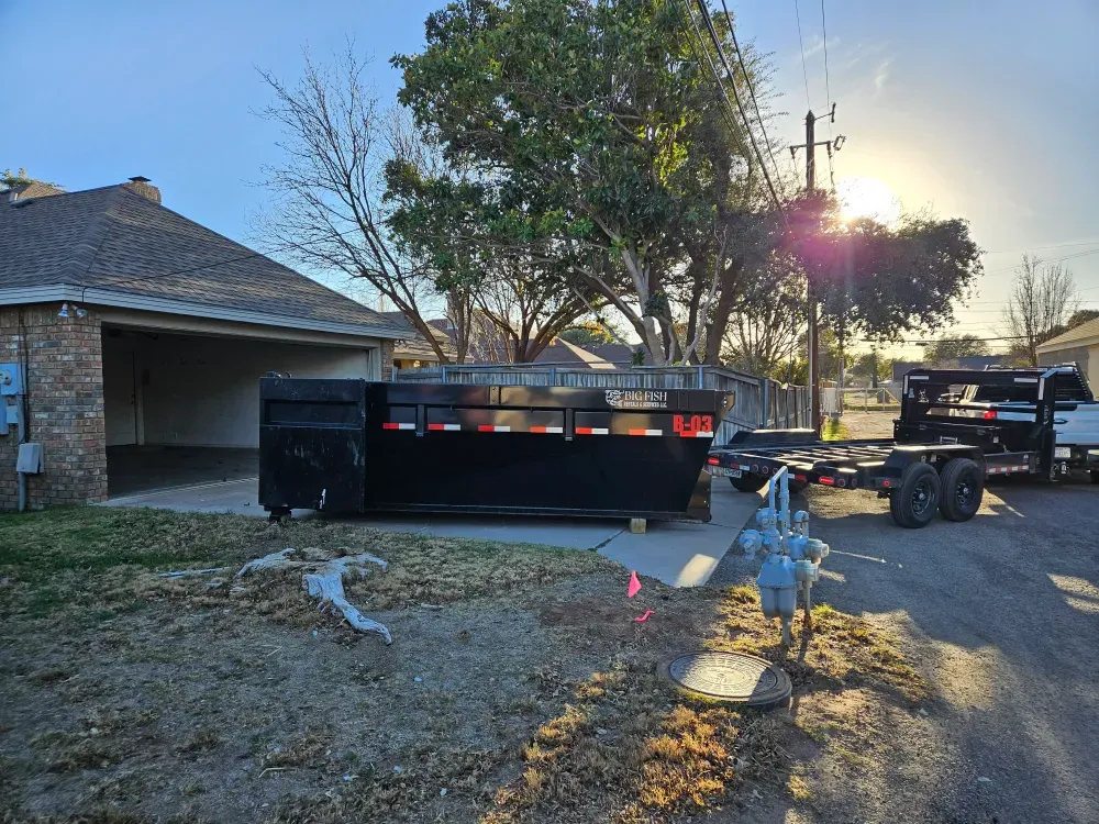 A Big Fish Rentals roll-off container set on a residential driveway in Pecos, Texas at golden hour, the hooklift trailer parked at the kerb and sun flaring through a tree.