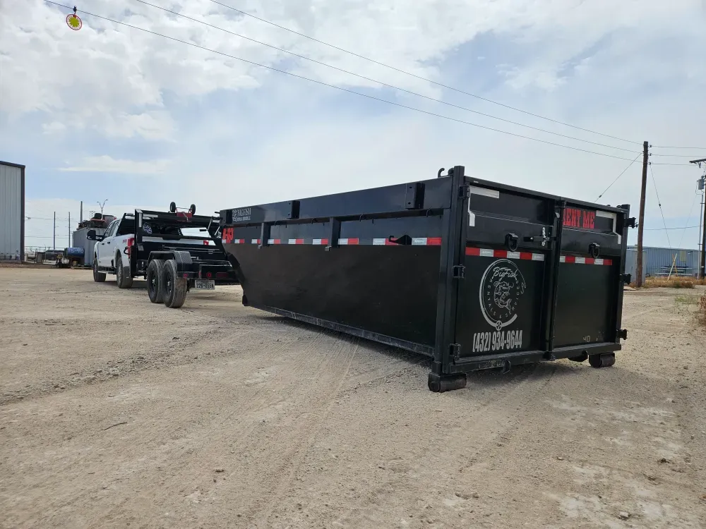Big Fish Rentals roll-off container freshly set on a caliche yard in Monahans, Texas, with the hook-lift trailer and the company pickup parked behind it under broken cloud.