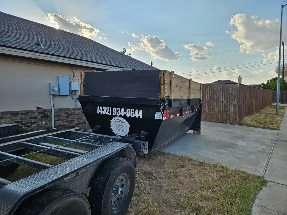A Big Fish Rentals roll-off container with wood side extensions standing on a home's concrete driveway in the evening, between the house and a cedar fence, with the hooklift trailer in the foreground.