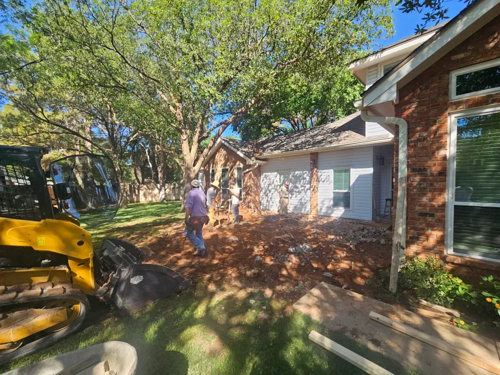 Broken concrete cleared from beside a brick house, with a tracked loader parked at the edge of the frame and three workers on the bare dirt where the slab came out.