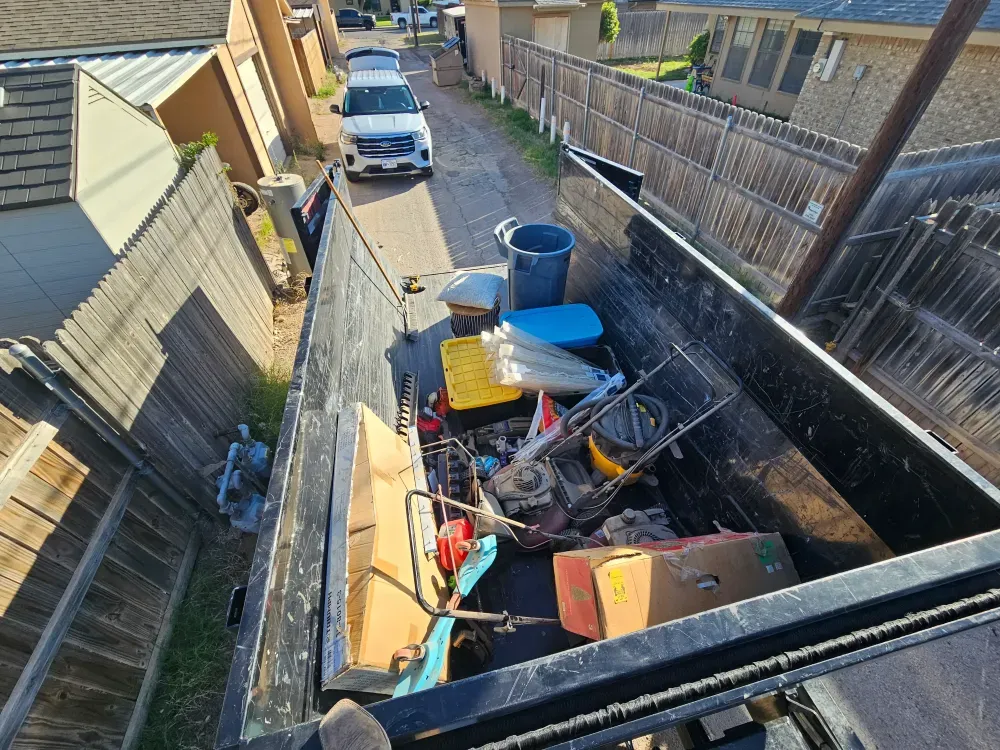 Big Fish Rentals roll-off container standing in a narrow alley in Monahans, Texas, part-loaded with a push mower, a trash can, a plastic tote and cardboard boxes from a house cleanout.