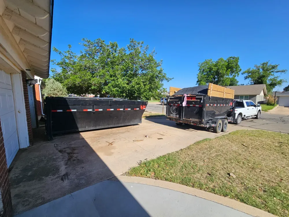 Low-sided black roll-off container parked on a concrete driveway beside a brick house, with the Big Fish Rentals pickup and loaded dump trailer waiting at the kerb in Odessa, Texas.
