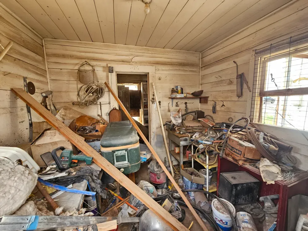 Inside a small wooden outbuilding packed floor to ceiling with stored junk, buckets, an old cooler, loose lumber and stacked boxes, waiting to be cleared out.
