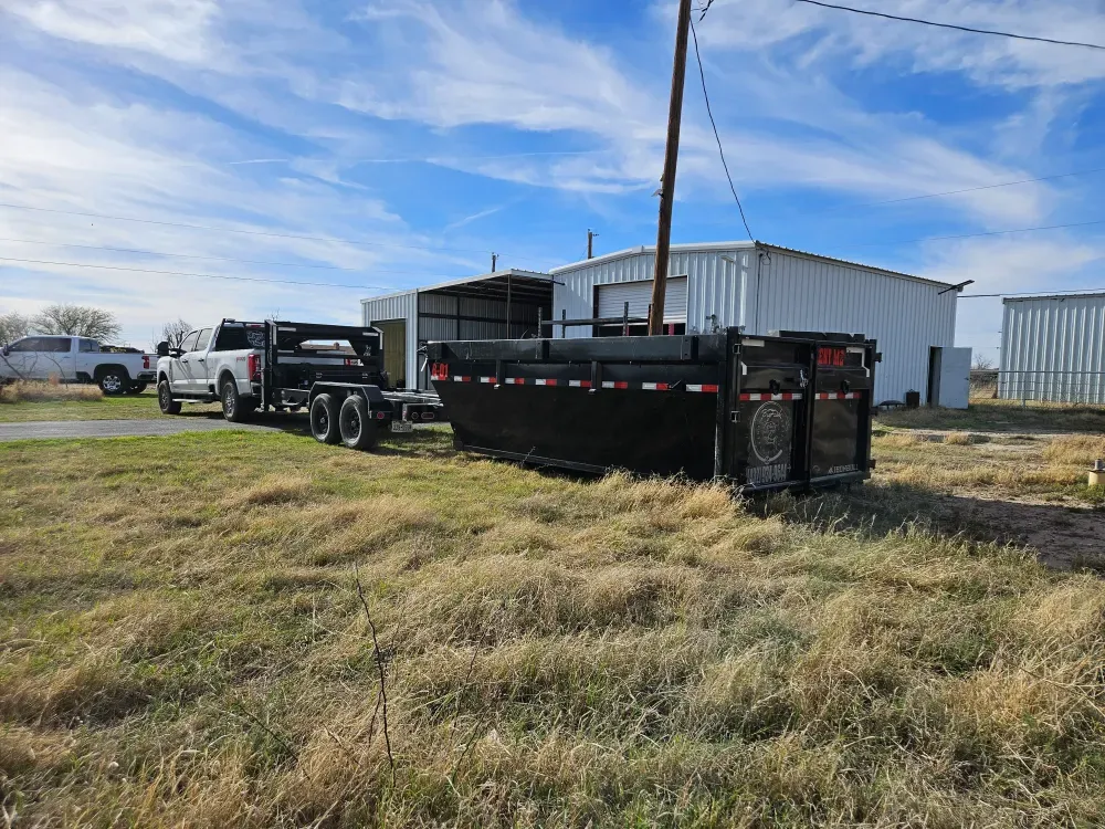 Roll-off container set down on dry winter grass next to a white metal shop building, with the Big Fish pickup and hook trailer parked behind it under a wide West Texas sky.