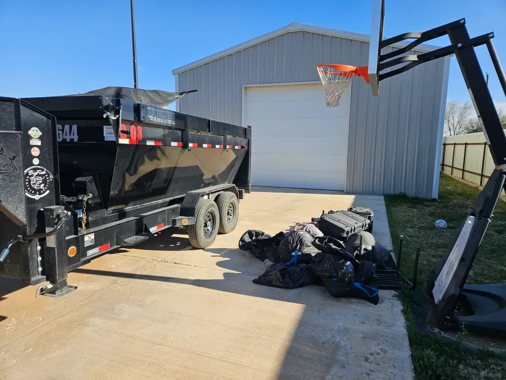 Big Fish Rentals dump trailer backed up a concrete driveway beside a row of filled black bags and a hard case waiting to be loaded, Odessa area.