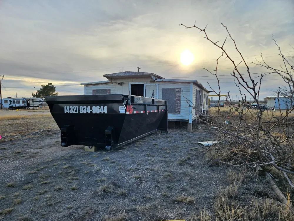 A Big Fish Rentals roll-off container standing on dry grass in front of a boarded-up house in Big Spring, Texas, sun low behind thin cloud.