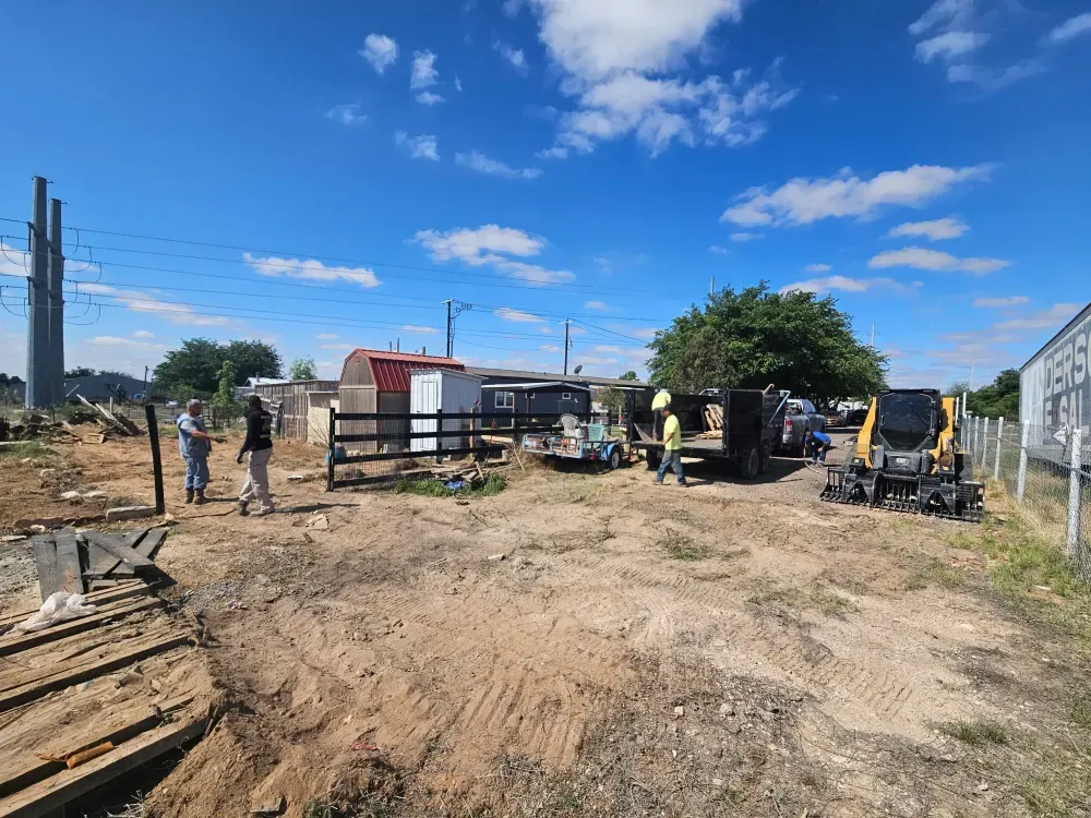 Crew clearing a lot near Midland, Texas: a track loader with an attachment mounted, men loading scrap lumber onto a trailer and stacked pallets in the foreground under big cumulus cloud.