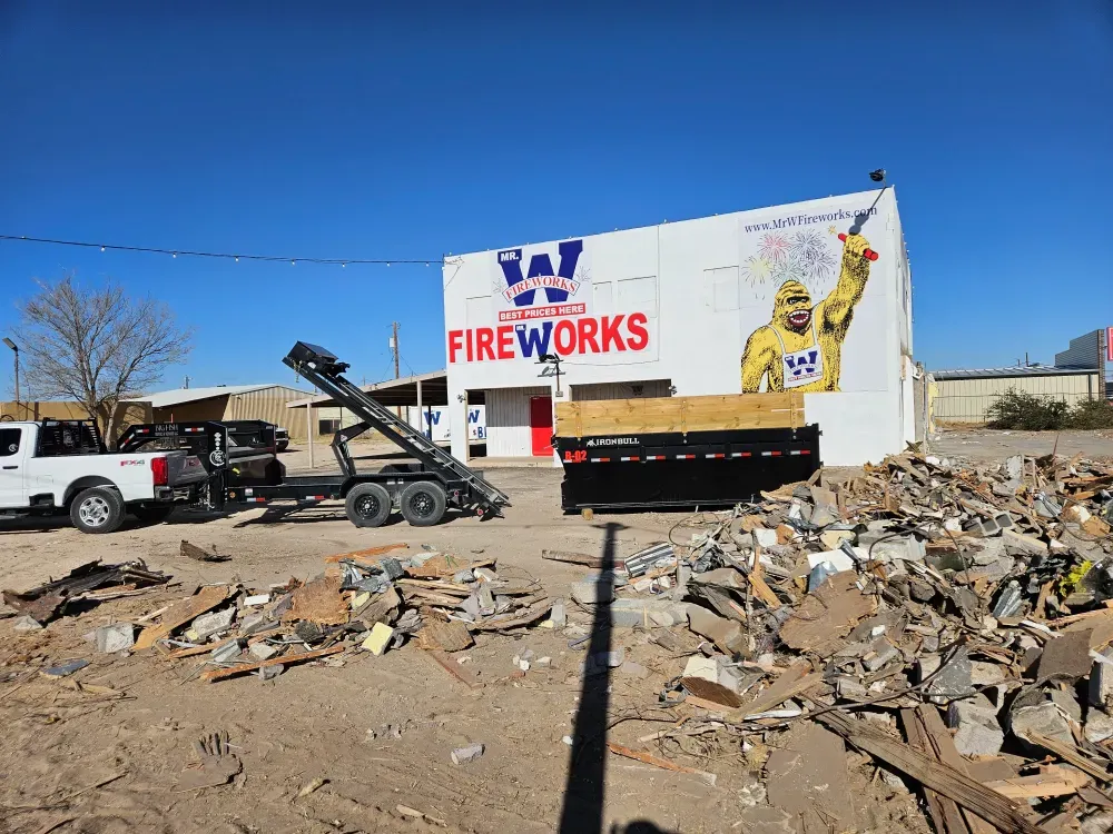 Big Fish Rentals roll-off container and hook-lift trailer set at a commercial building with a field of torn-out lumber, sheet metal and rubble across the foreground.