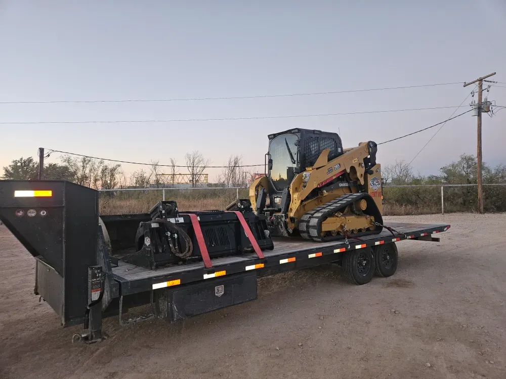 CAT compact track loader chained down on a gooseneck flatbed trailer at dusk with spare attachments loaded ahead of it, ready to move to a job site near West Odessa, Texas.