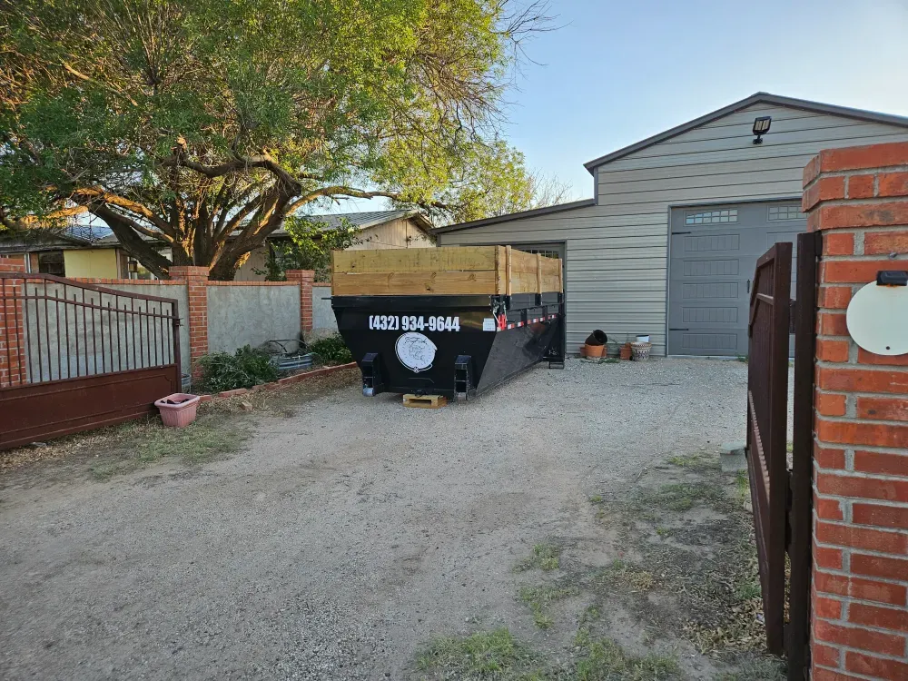 A Big Fish Rentals container set down on the gravel drive beside a detached garage in West Odessa, Texas, brick gate posts open and the garage door shut.