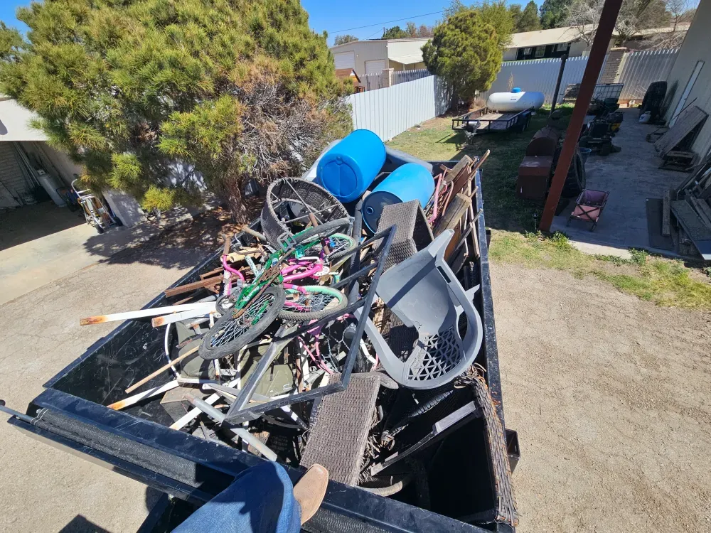 Looking down into a Big Fish container loaded with a household cleanout: children's bikes, patio chairs, blue barrels and broken furniture, parked in the yard beside the house.