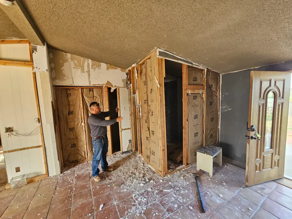 A worker pulling ruined wall board off the studs inside a vacant house, debris scattered across the terracotta tile floor and the front door open to daylight.