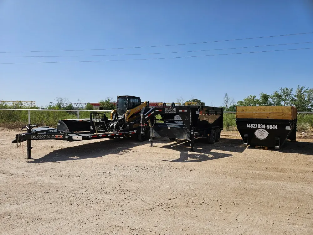 Big Fish Rentals equipment lined up on a caliche yard in West Texas: a compact track loader on an equipment trailer, the gooseneck dump trailer, and a roll-off container with timber side extensions.