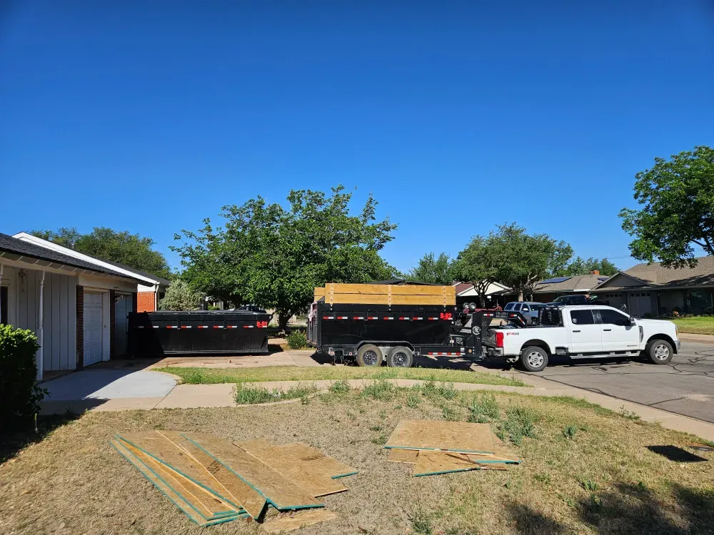 Roll-off container parked at the garage on a remodel job, the Big Fish truck and dump trailer at the kerb, sheets of OSB and framing lumber laid out on the front lawn.