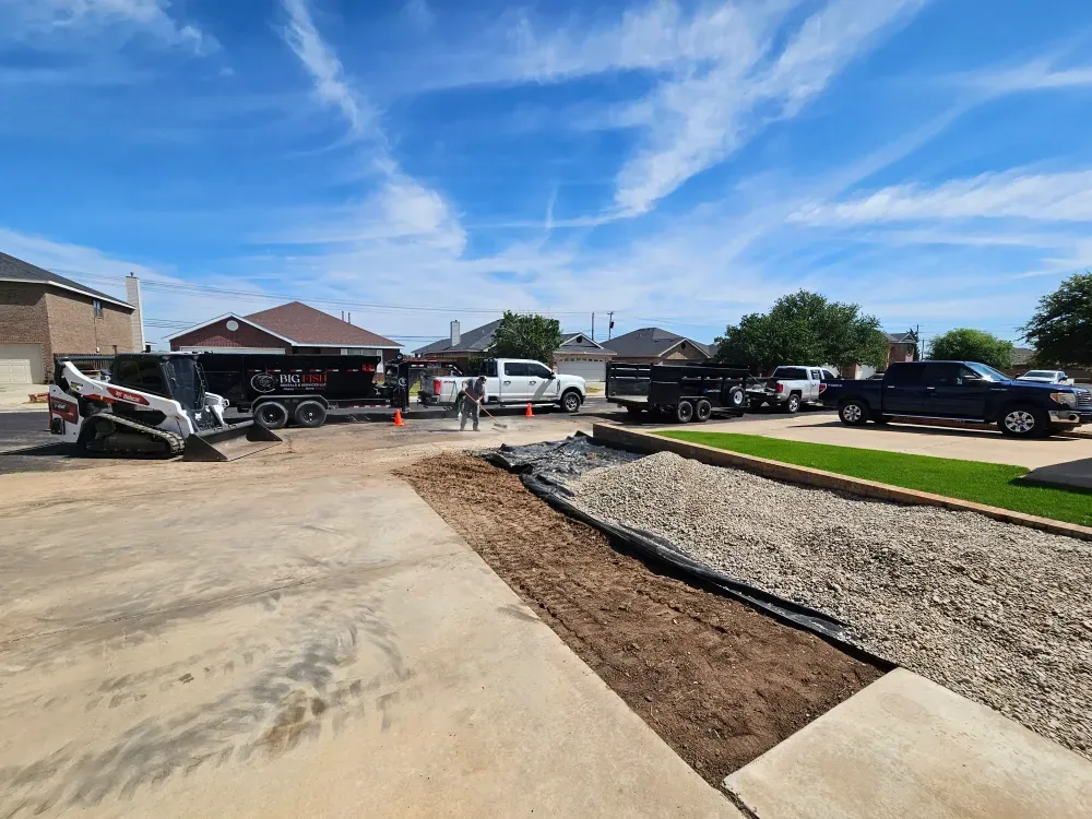 Compact track loader parked on a fresh concrete driveway while a worker rakes a new gravel bed, the Big Fish Rentals dump trailer and truck waiting at the kerb in West Odessa, Texas.