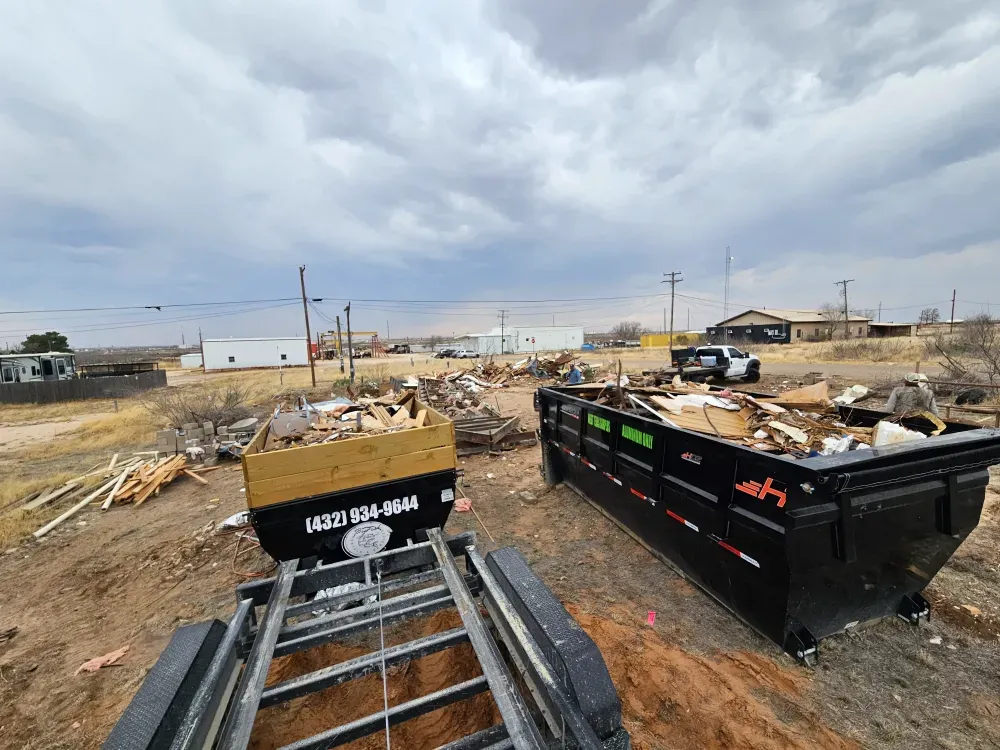 Big Fish Rentals roll-off container filled to the top rail with demolition debris — broken lumber, drywall and siding — on a job site under a stormy West Texas sky, with another contractor's container behind it.