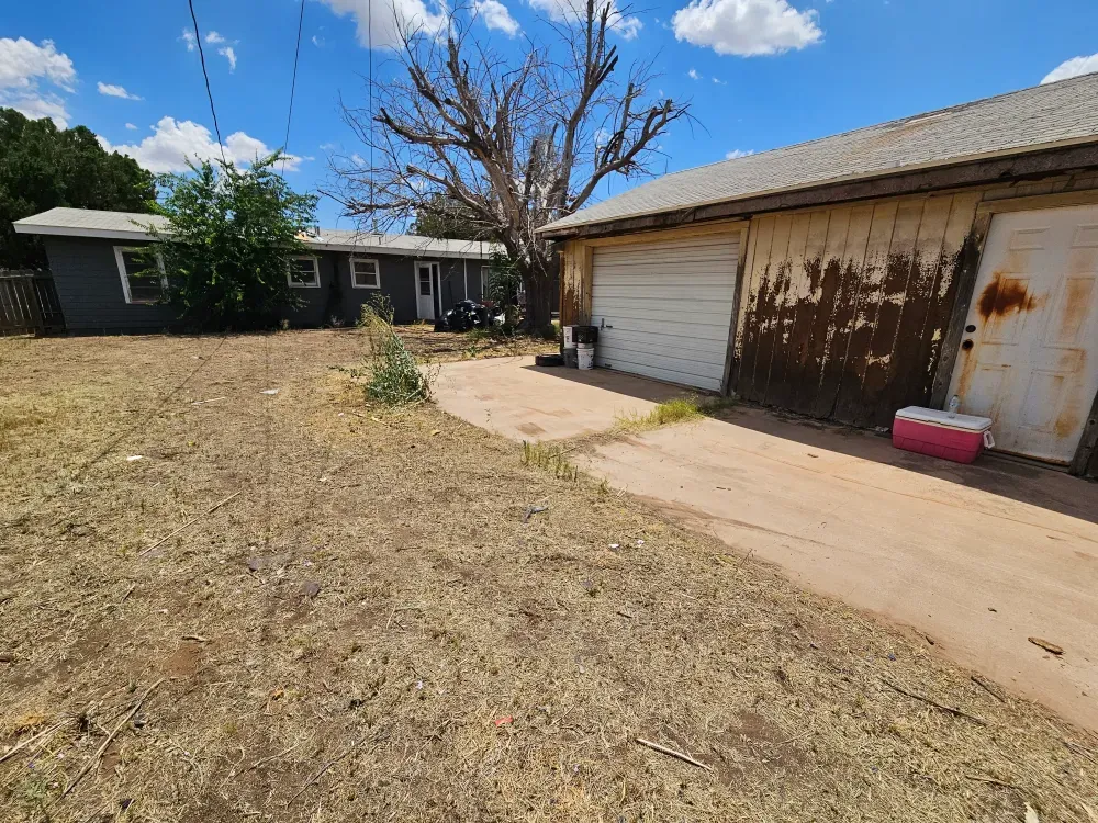 The same backyard after Big Fish Rentals hauled everything out: the concrete apron swept clear and the ground raked, with the same rusted garage and pink cooler still in frame.