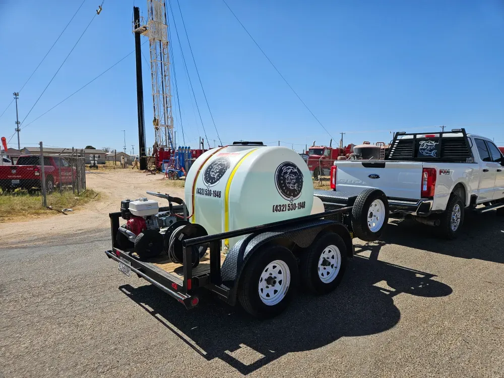 Trailer mounted pressure washer with a white poly water tank carrying the Big Fish Rentals logo, a Honda engine pump and a hose reel, hitched to the pickup on an oilfield yard with a workover rig behind.