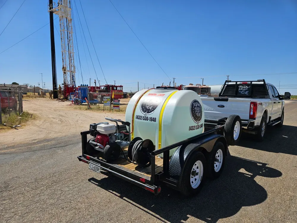 Rear view of the pressure washer trailer on a paved lease road, the white tank marked RENTAL UNIT with the pump and hose reel stowed, a workover rig standing behind it in West Texas.