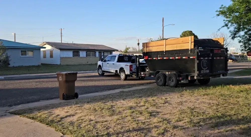 The Big Fish Rentals truck at the kerb of a Kermit, Texas street at golden hour, towing a dump trailer with tall wood side extensions and the phone number on the side.