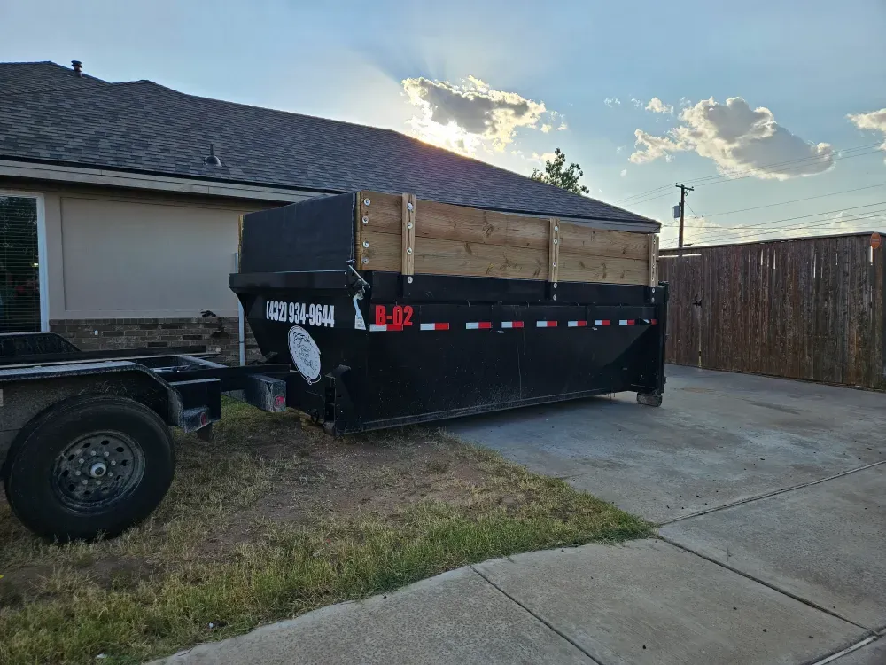 Roll-off container B-02 with timber side extensions and the (432) 934-9644 number set on a residential concrete driveway at sunset, the hook-lift trailer alongside and room left to walk past it.