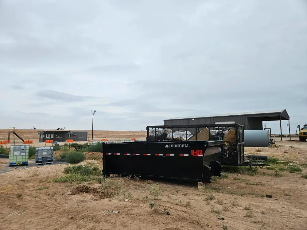Big Fish Rentals container B-03 on a remote lease location, with a caged trailer of bagged site waste, IBC totes and a metal building behind it under grey West Texas sky.