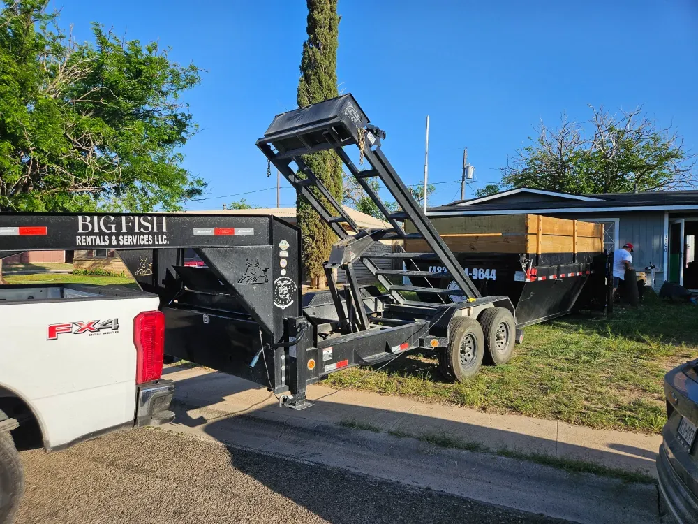 Big Fish hook trailer with its arm raised, BIG FISH RENTALS & SERVICES LLC lettered across the gooseneck, having just set a roll-off container on the grass in front of a house while a worker in a red cap stands alongside in golden evening light.