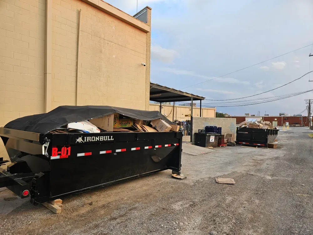 Container B-01 on timber blocks in the alley behind a commercial building, tarp pulled back and the box heaped with flattened cardboard, a second full container further down the alley.