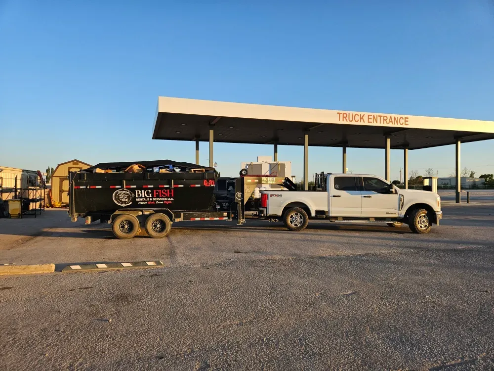 Big Fish Rentals truck at golden hour towing the tarped, fully loaded dump trailer under the TRUCK ENTRANCE canopy at the disposal site, company name and tagline readable on the side.