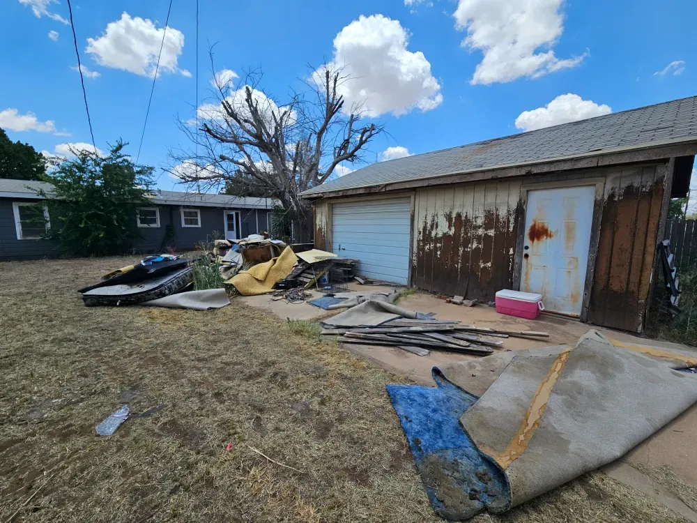 Backyard of a West Texas house before the cleanout: soiled carpet rolled out across the concrete apron, torn-out lumber and an old jet ski hull on the dead lawn beside a rusted metal garage.